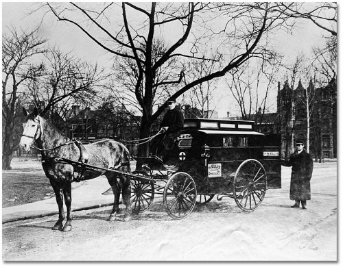 A small covered carriage pulled by one horse, with a man sitting on the front driving, and another standing at the back by the open back door.