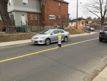 Image of a residential street with 1 lane of traffic going each way. On the painted yellow centre line, there is a in road flexible speed sign that's 3 feet high and reads maximum of 40 km/h.