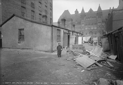 Black and white photograph of a young girl standing outside a one storey building with Toronto's old City Hall in the background.