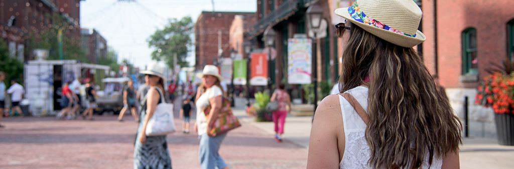 Back of a woman in a hat looking towards an area with shops and other shoppers.