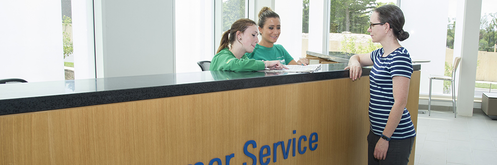 Two City staff sit behind a Customer Service desk and speak with a visitor at a community recreation centre.