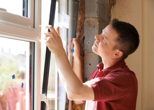 A man wearing a red shirt, installing a window in a home.