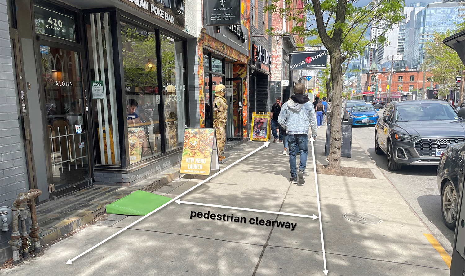 A city sidewalk scene with a temporary green accessibility ramp positioned at the entrance of a storefront. Two white arrows overlaid on the image show the edges of the pedestrian clearway, a direct, unobstructed path between the ramp and the tree line near the curb. The image illustrates how the ramp should be placed to maintain the clearway for pedestrian movement.
