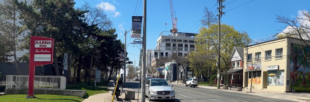 Photo with view facing west on Dundas Street West showing buildings with storefronts, street trees, and signage on the north and south sides of the street, a mural on the side main wall of one of the buildings on the north side of the street, and a mid-rise building under construction.